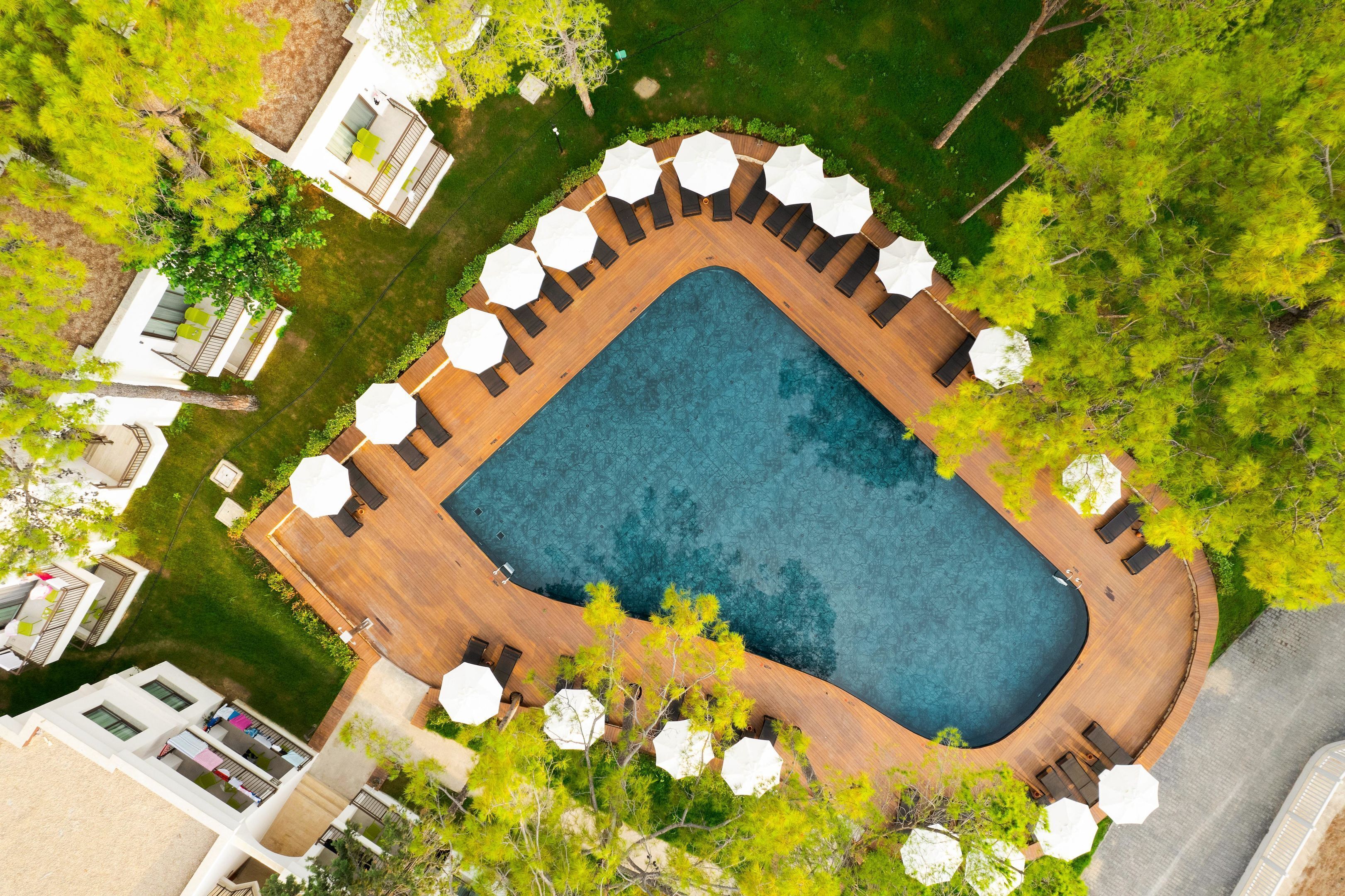 Vista aérea de una piscina con muelle de madera, rodeada de sillones de descanso y sombrillas, con edificios y árboles al fondo.