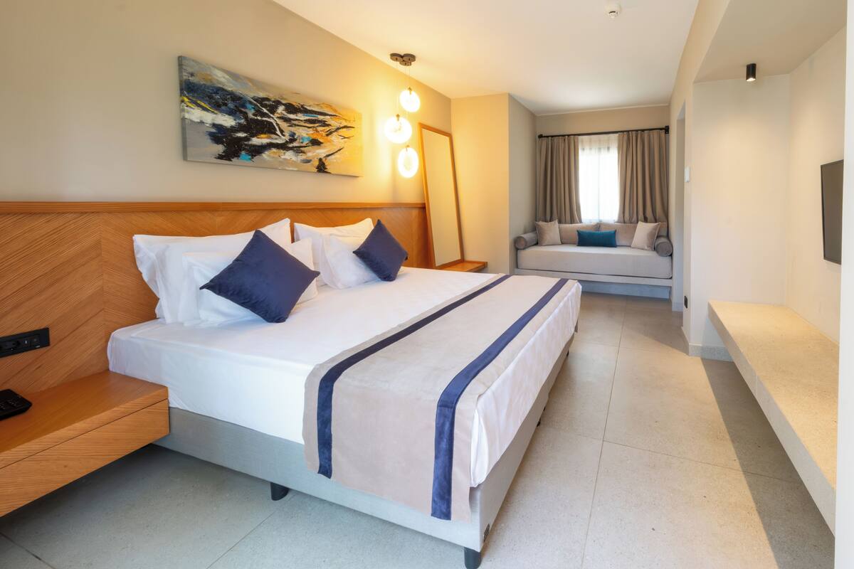 A hotel room with a large bed, a bedside table, a chair, a sofa, a painting on the wall, and a window with curtains.