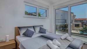 Un dormitorio moderno con una cama grande, dos ventanas, un balcón y vistas a un edificio y piscina.