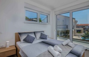 Une chambre moderne avec un grand lit, deux fenêtres, un balcon et une vue sur un immeuble et une piscine.