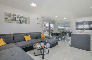 Un salon moderne avec un canapé d’angle gris, une table basse ronde sur laquelle sont disposés des fruits et des boissons, et un téléviseur à écran plat fixé au mur.