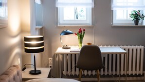 A hotel room with a bed, a desk with a lamp, a chair, a radiator, and a window with a plant.