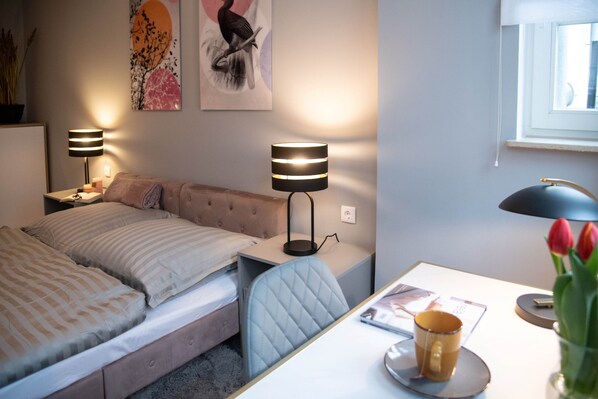 Un dormitorio con cama, mesitas de noche, lámparas, un escritorio, una silla, un ventanal y un jarrón con tulipanes.