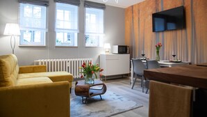 A modern living room with a yellow sofa, a wooden coffee table with a vase of flowers, a dining area with a table and chairs, and a flat-screen TV mounted on the wall.
