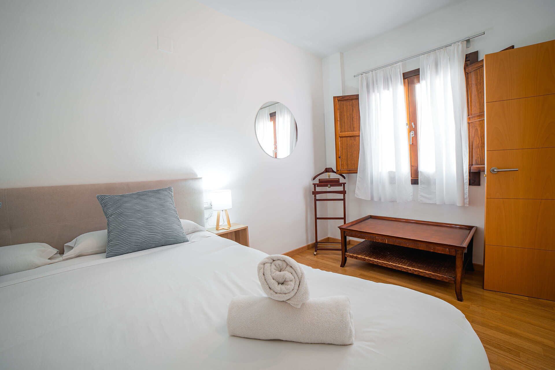 Una cama bien hecha con una toalla enrollada, una lámpara de noche y un banco de madera.