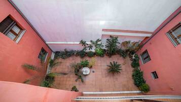Un estrecho pasillo con paredes rosadas y suelo de baldosas que conduce a una escalera. Las paredes están decoradas con diversas plantas en macetas, y hay ventanas con contraventanas de madera a ambos lados.