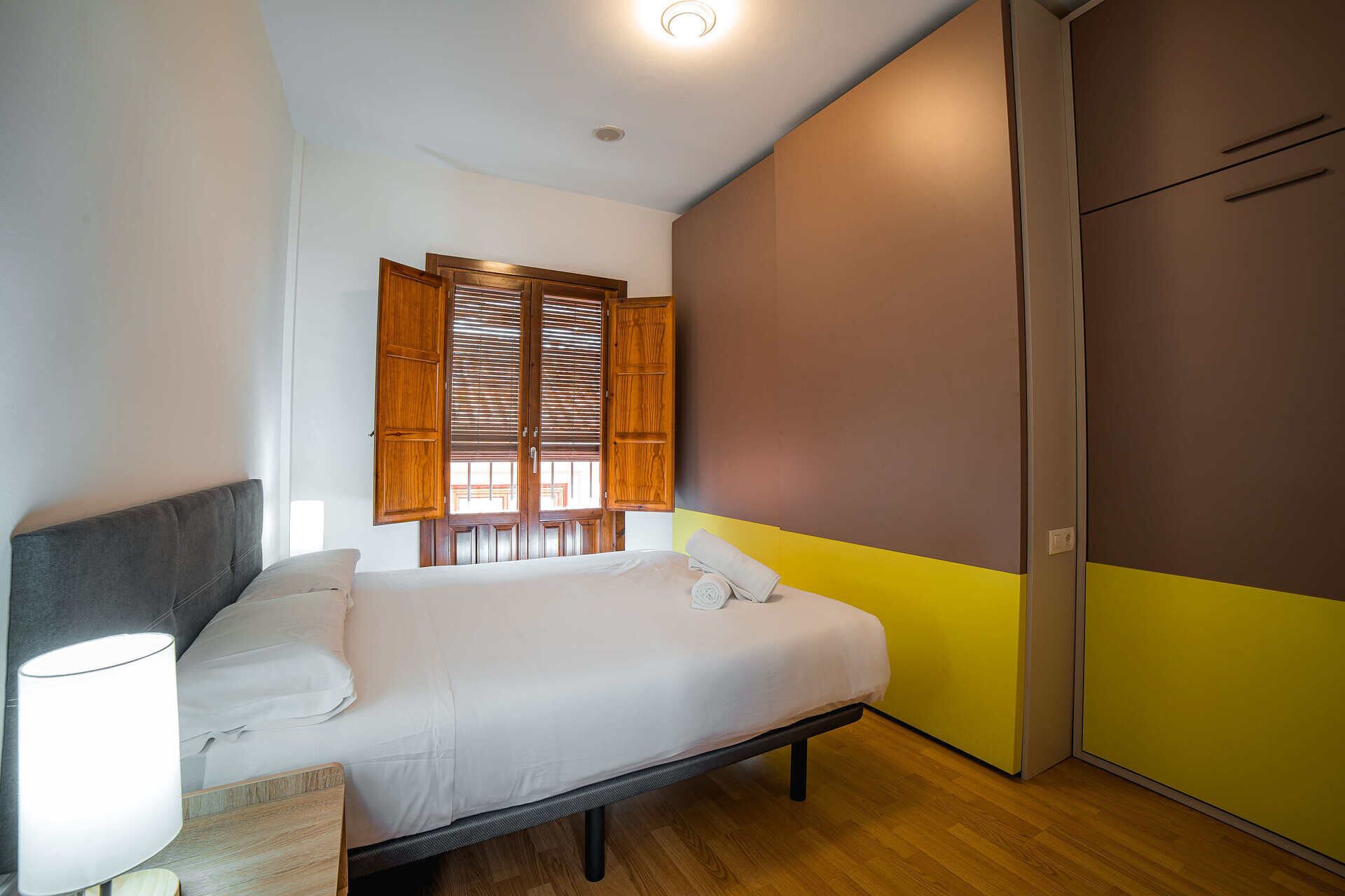 Une chambre à coucher comprenant un lit, une table de chevet, une lampe, une fenêtre avec des volets en bois et une armoire.