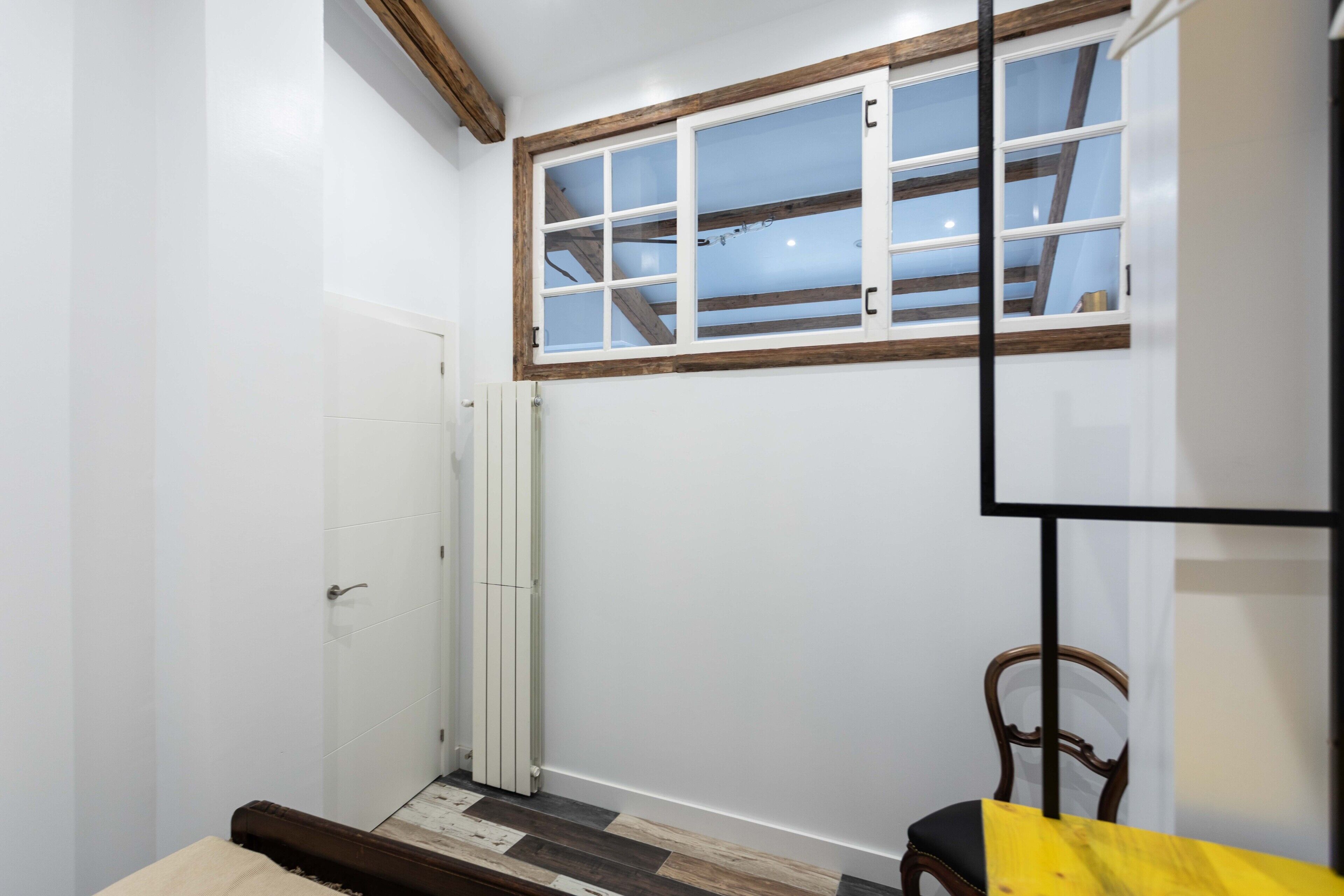 Une pièce avec une fenêtre à cadre en bois, une porte blanche, un radiateur, un plancher en bois et une table jaune.