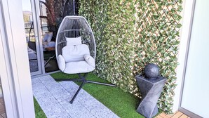 Een hangstoel met kussen, een kleine stenen plantenbak en een groene muur met klimplanten.