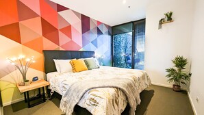 Een moderne slaapkamer met een wand met een geometrisch patroon, een groot bed met veel kussens, een nachtkastje met een lamp, en uitzicht op bomen door het raam.