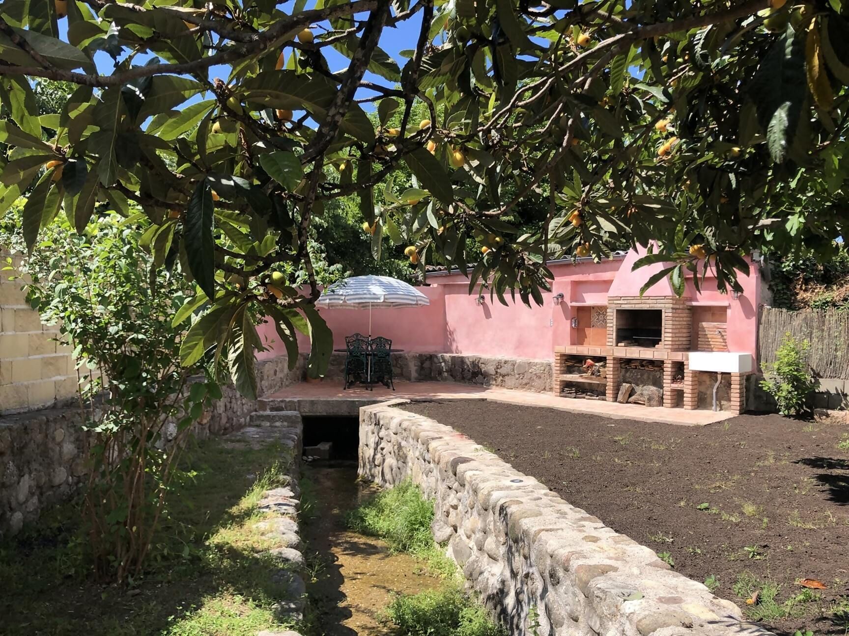 Un patio con un muro de piedra, un horno de ladrillo y una mesa con sillas bajo un árbol.