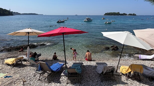 A pebbled beach with lounge chairs and umbrellas, a clear blue sea, and boats in the distance.