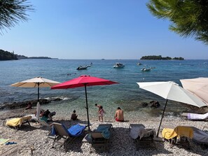 Una playa de guijarros con sillas de playa y sombrillas, un mar azul cristalino y barcos a lo lejos.