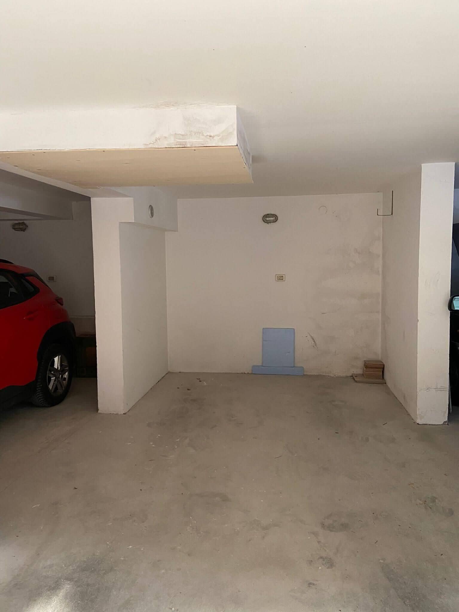 Un garage con un’auto rossa parcheggiata all'interno, pavimento in cemento e pareti bianche.