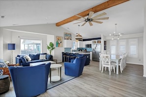 A spacious living room with a sofa set, a dining table with chairs, a kitchen area, and a ceiling fan.