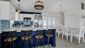 A modern kitchen with a central island, bar stools, white cabinetry, and a dining area with white chairs. - Oceanfront Bliss - Spacious Beach Cottage for up to 11 Guests (Surf City)