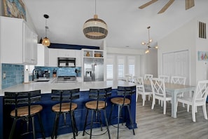 A modern kitchen with a central island, bar stools, white cabinetry, and a dining area with white chairs.