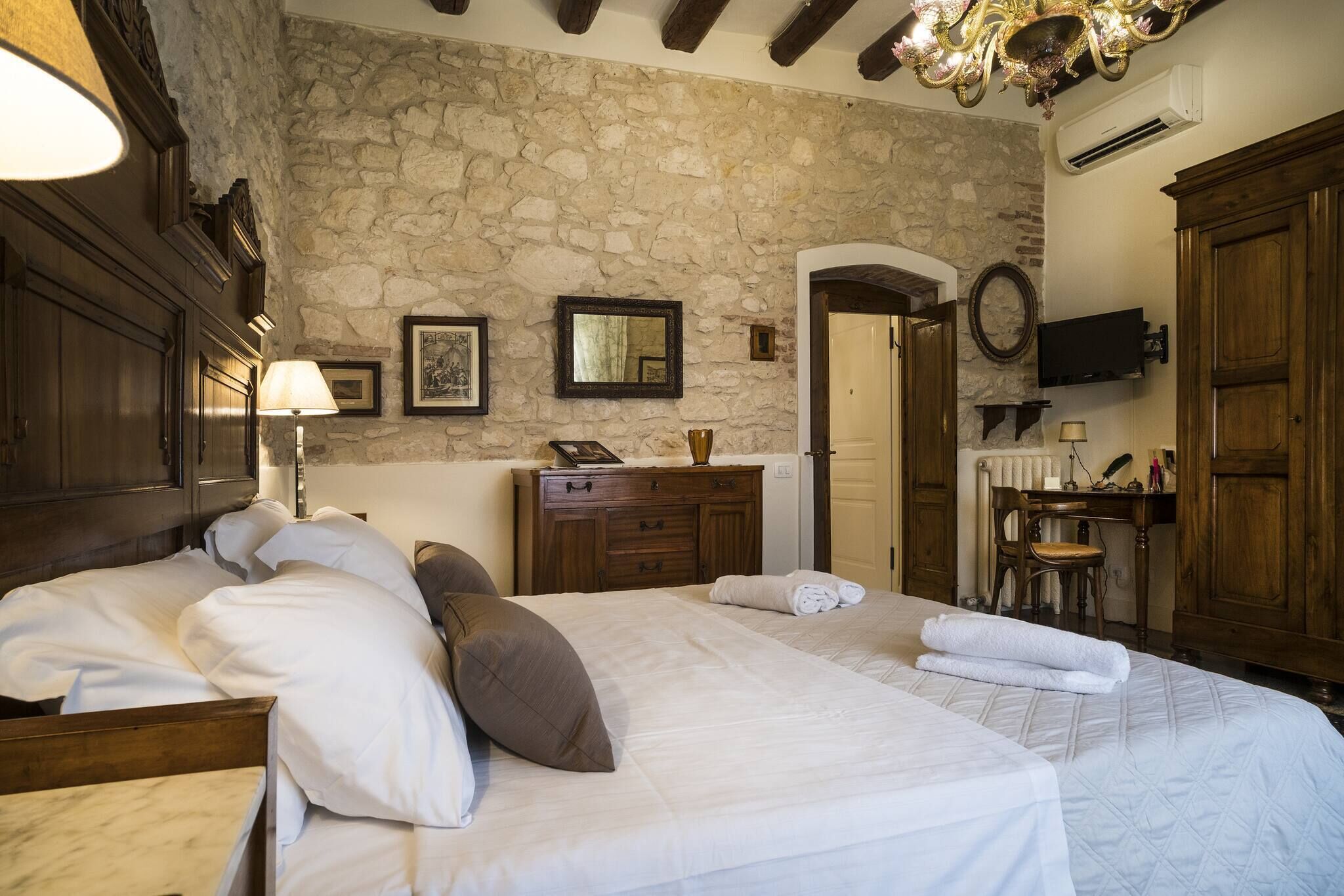 A bedroom with a stone wall, wooden furniture, a bed with pillows, a dresser, a mirror, a television, and a chandelier.