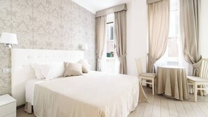 Une chambre d’hôtel avec un grand lit, une table de chevet avec une lampe, une chaise, une petite table recouverte d’une nappe et une fenêtre avec des rideaux.