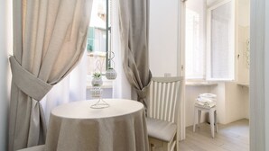 Une pièce avec une table ronde, une chaise, une fenêtre avec des rideaux, un petit tabouret avec des serviettes et une plante sur le rebord de la fenêtre.