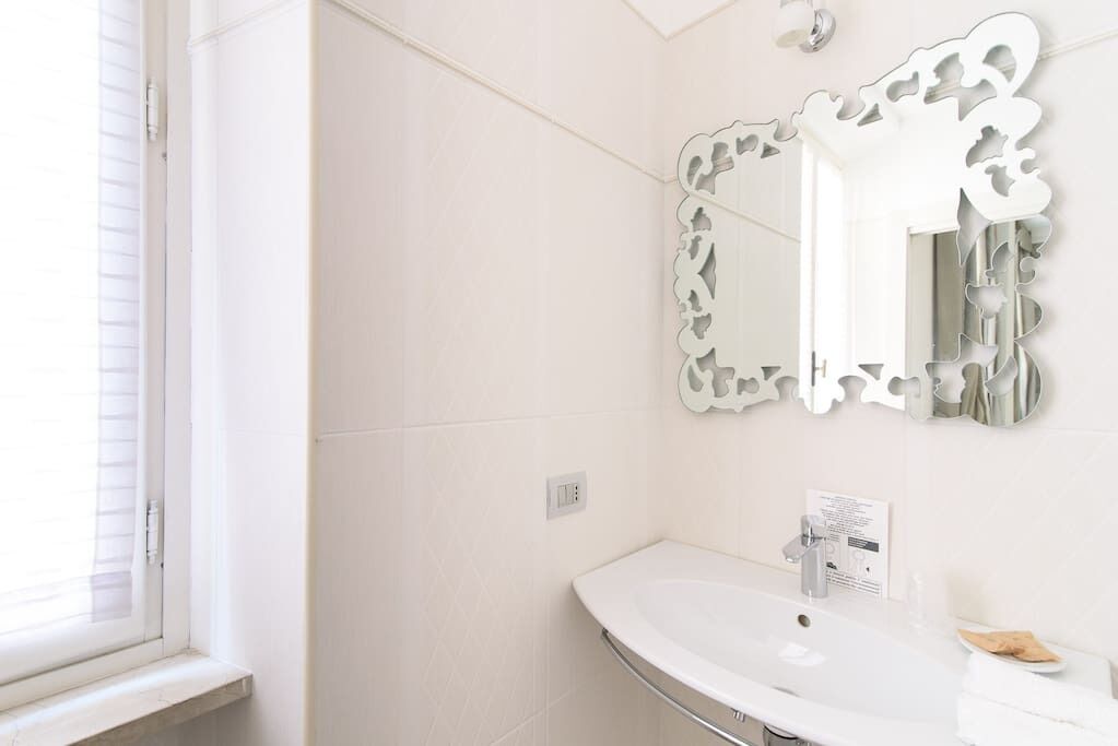 A bathroom with a white sink, a mirror with a decorative frame, and a window with blinds.