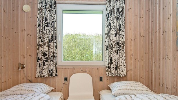 Une chambre avec deux lits simples, une chaise blanche, des murs en bois, une fenêtre avec des rideaux et une vue sur la verdure à l’extérieur.