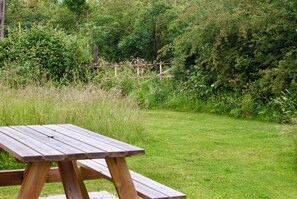 A wooden picnic table in a grassy area with dense shrubbery and trees in the background. - Light and airy stylish barn conversion set in peaceful countryside. (Henley in arden)