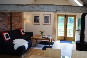 A living room with a brick wall, a dark sofa with a white throw, a wooden coffee table with books and a vase of flowers, and two framed pictures on the wall. - Light and airy stylish barn conversion set in peaceful countryside. (Henley in arden)