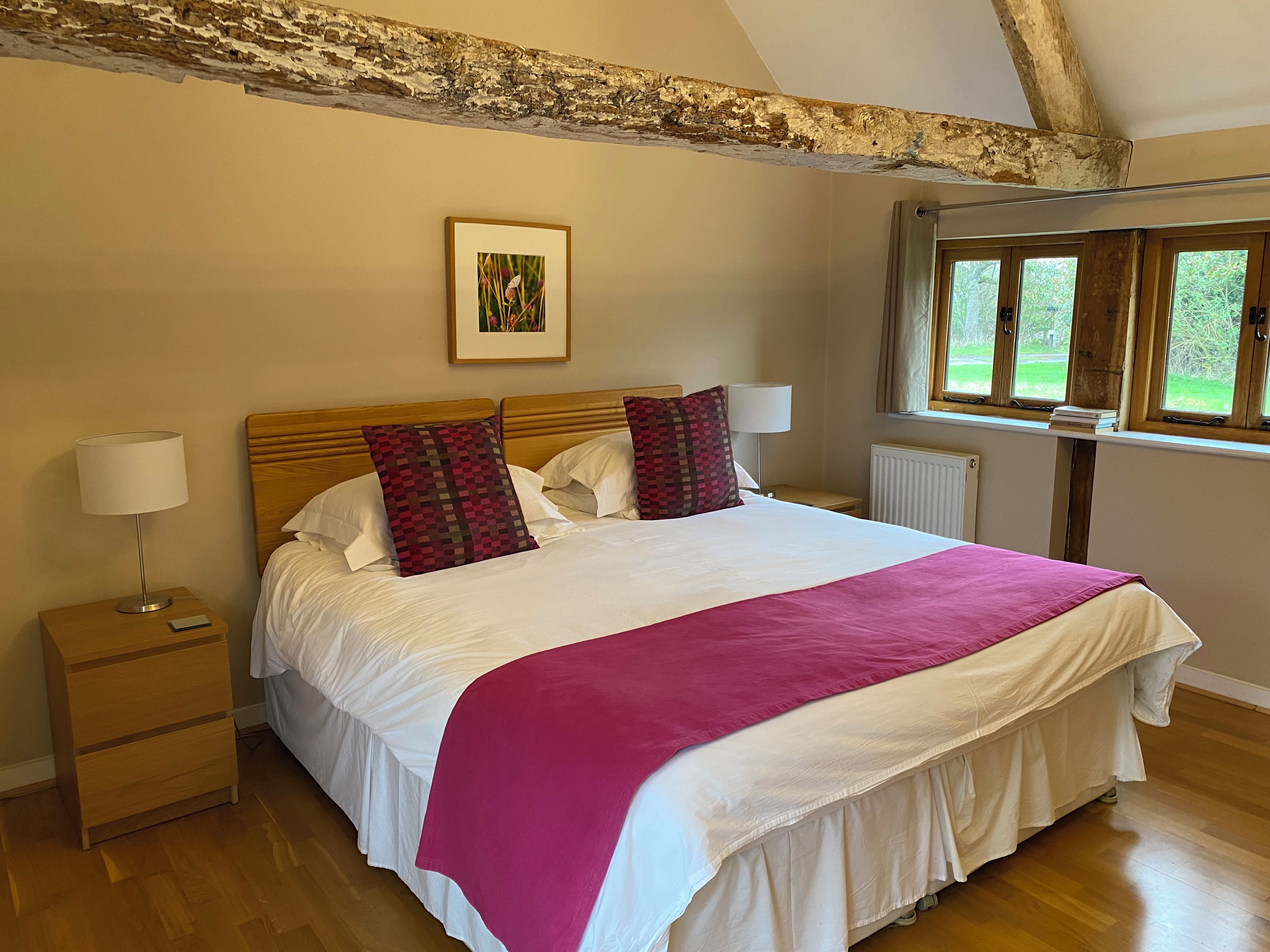 A bedroom with a wooden bed, white bedding, a pink throw, a nightstand with a lamp, and a window with a view of greenery.