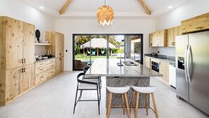 A modern kitchen with wooden cabinets, a central island with stools, a stainless steel refrigerator, and a view of a tropical outdoor area. - 5 Min Drive to the Beach, Private Heated Jacuzzi - Sleeps 20 (Playa Avellana)