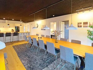 A modern dining area with a long wooden table, grey chairs, pendant lighting, and a vase with flowers. - 14 Person Holiday Home in Vaeggerlose (Vaeggerlose)