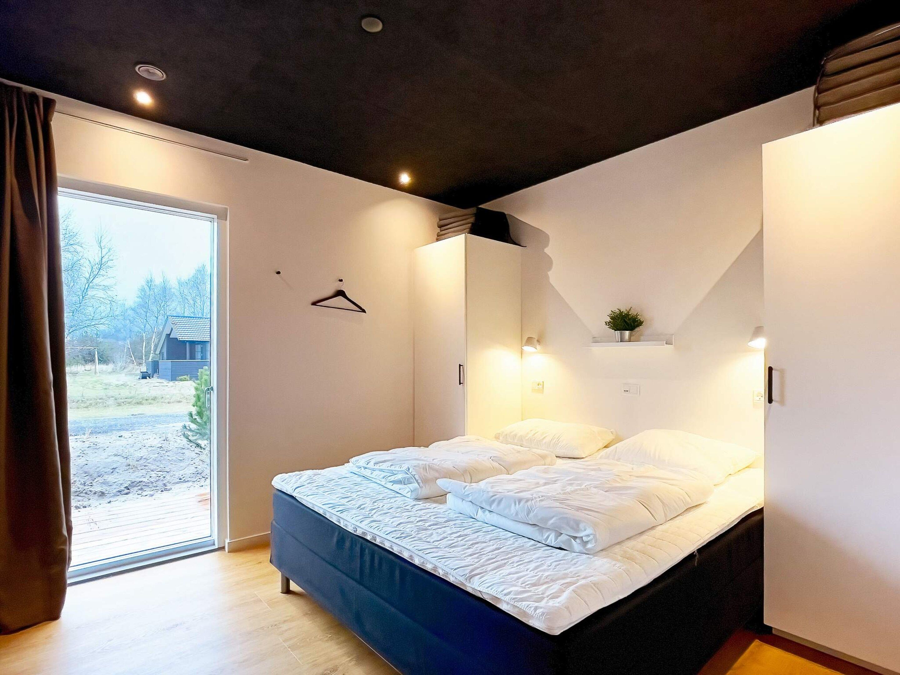 A bedroom with a bed, a hanging light, a plant on a shelf, and a view through a door.