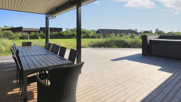 An outdoor patio with a wooden table and chairs, a covered area, and a view of buildings and greenery in the background. - 14 Person Holiday Home in Vaeggerlose (Vaeggerlose)
