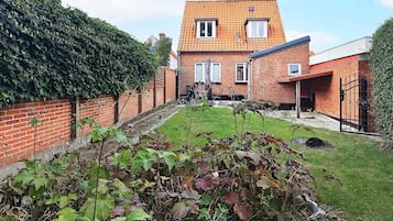 A brick house with a tiled roof, a garden with various plants, and a clear sky.