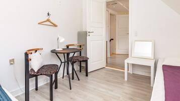 A compact room with a bed, a small table with a lamp, two chairs, and a hanging coat rack.