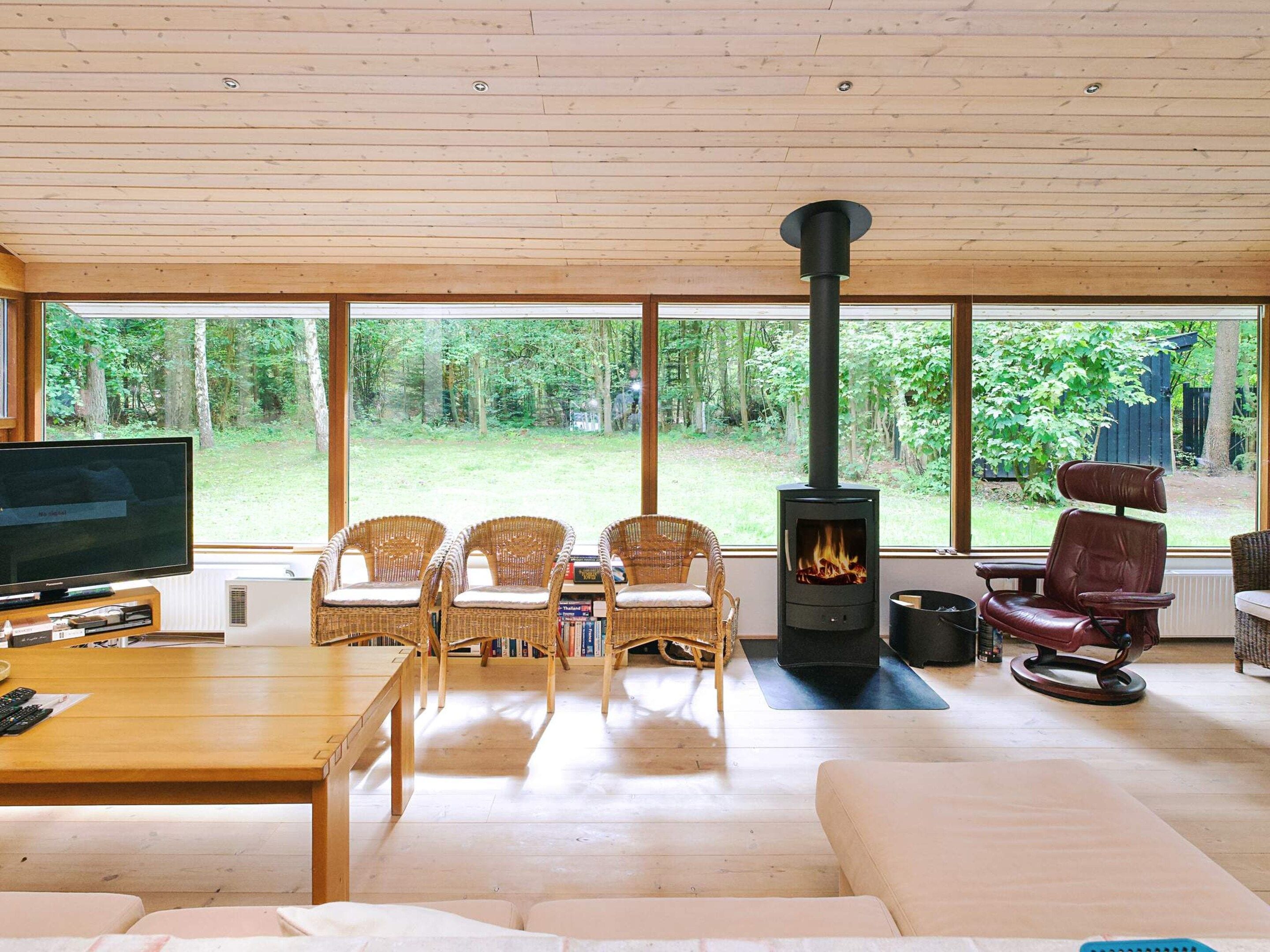 A cozy living room with a large TV, wicker chairs, a sofa, a fireplace, and a view of a wooded area.