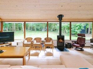 Un salon confortable avec une grande télévision, des fauteuils en osier, un canapé, une cheminée et une vue sur une zone boisée.