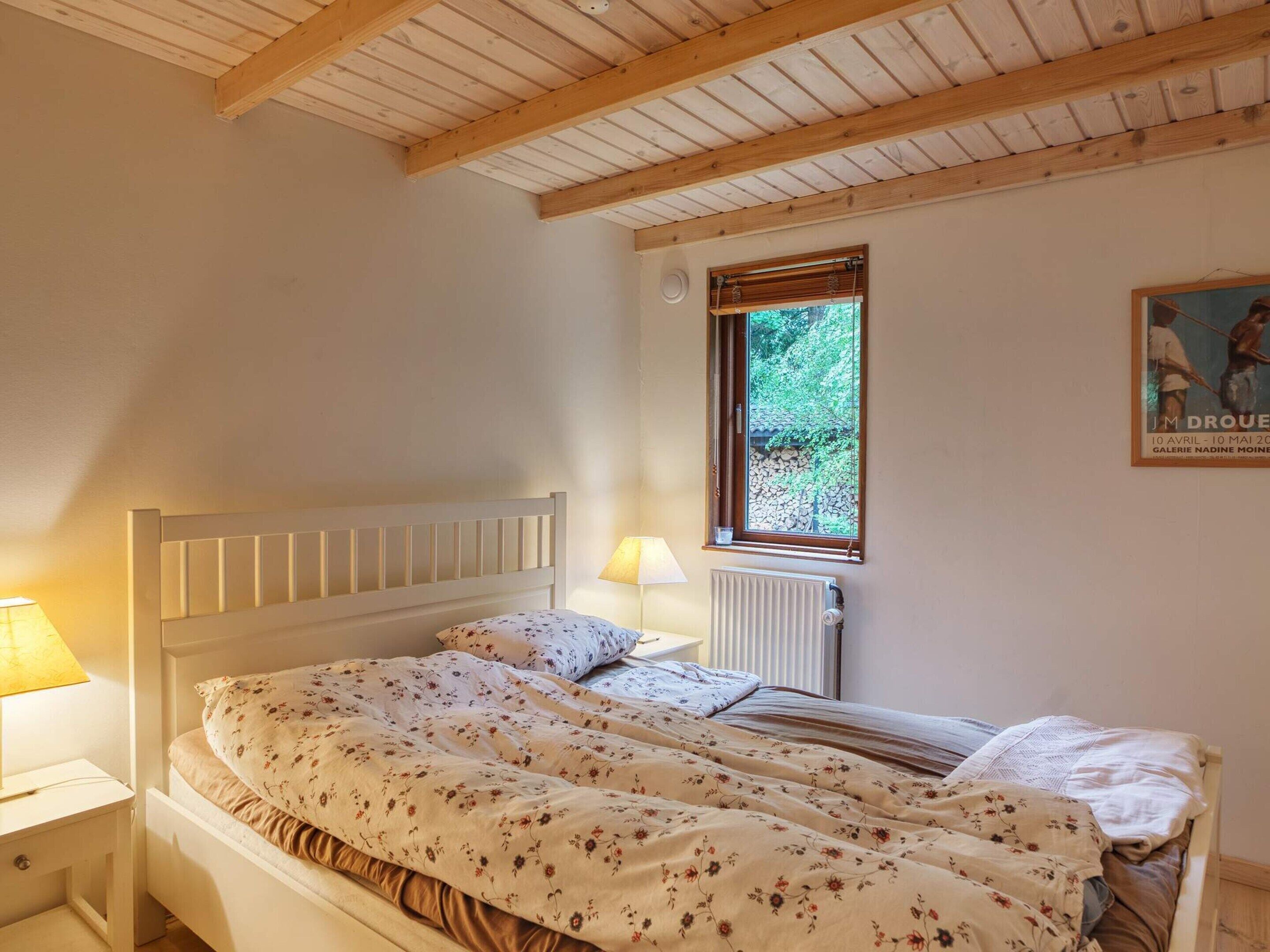 A bedroom with a wooden ceiling, a single bed with floral bedding, a window with a view of greenery, and a poster on the wall.