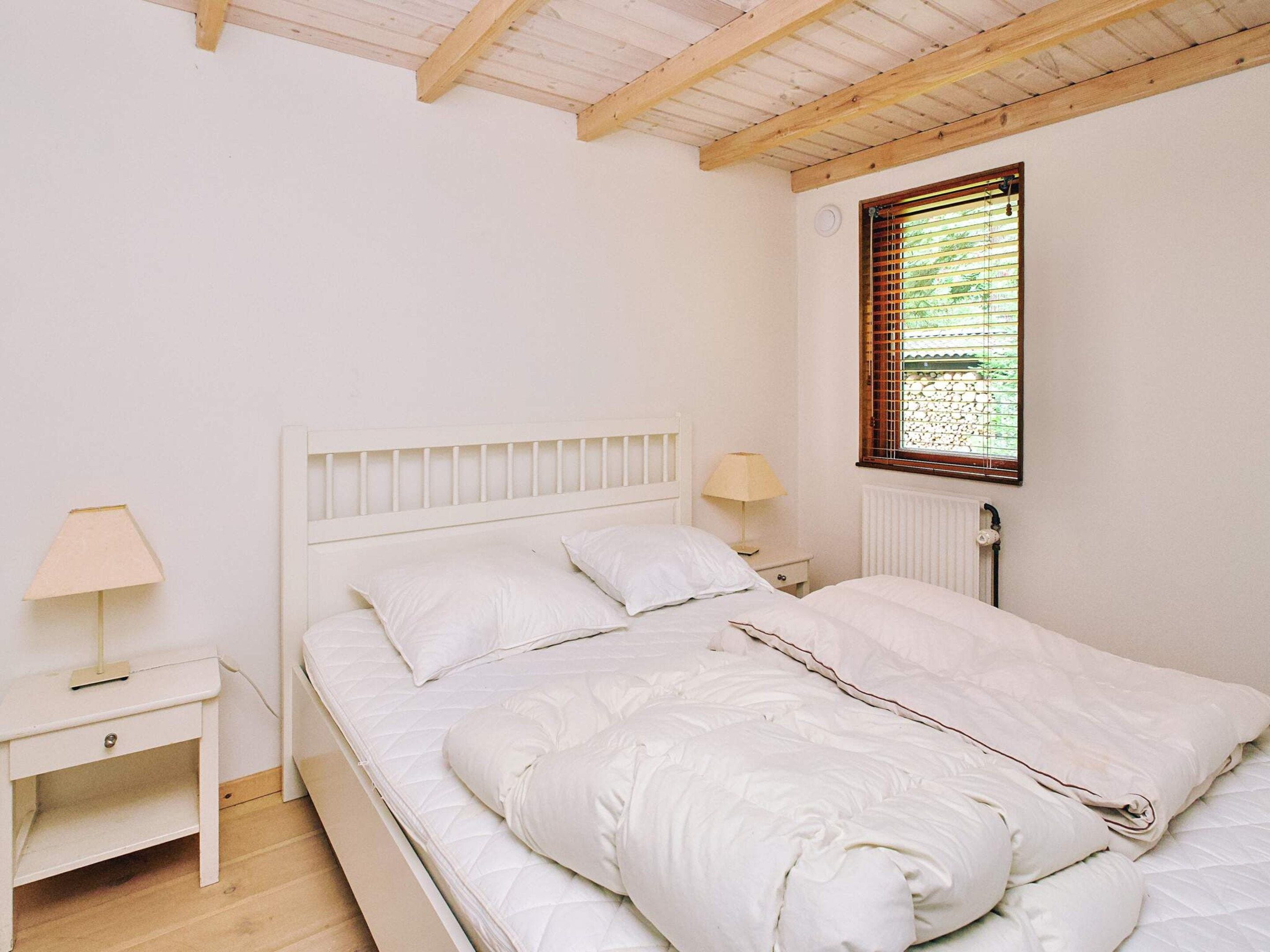 Ein Schlafzimmer mit einem weißen Bett, einem Nachttisch, einer Lampe und einem Fenster mit Jalousien.