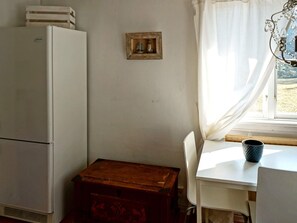 A compact room with a white refrigerator, a wooden chest, a white table, and a window with curtains.
