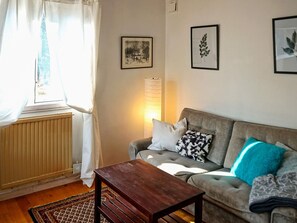 A living room with a sofa, a wooden coffee table, a radiator, a window with curtains, and wall-mounted artwork.