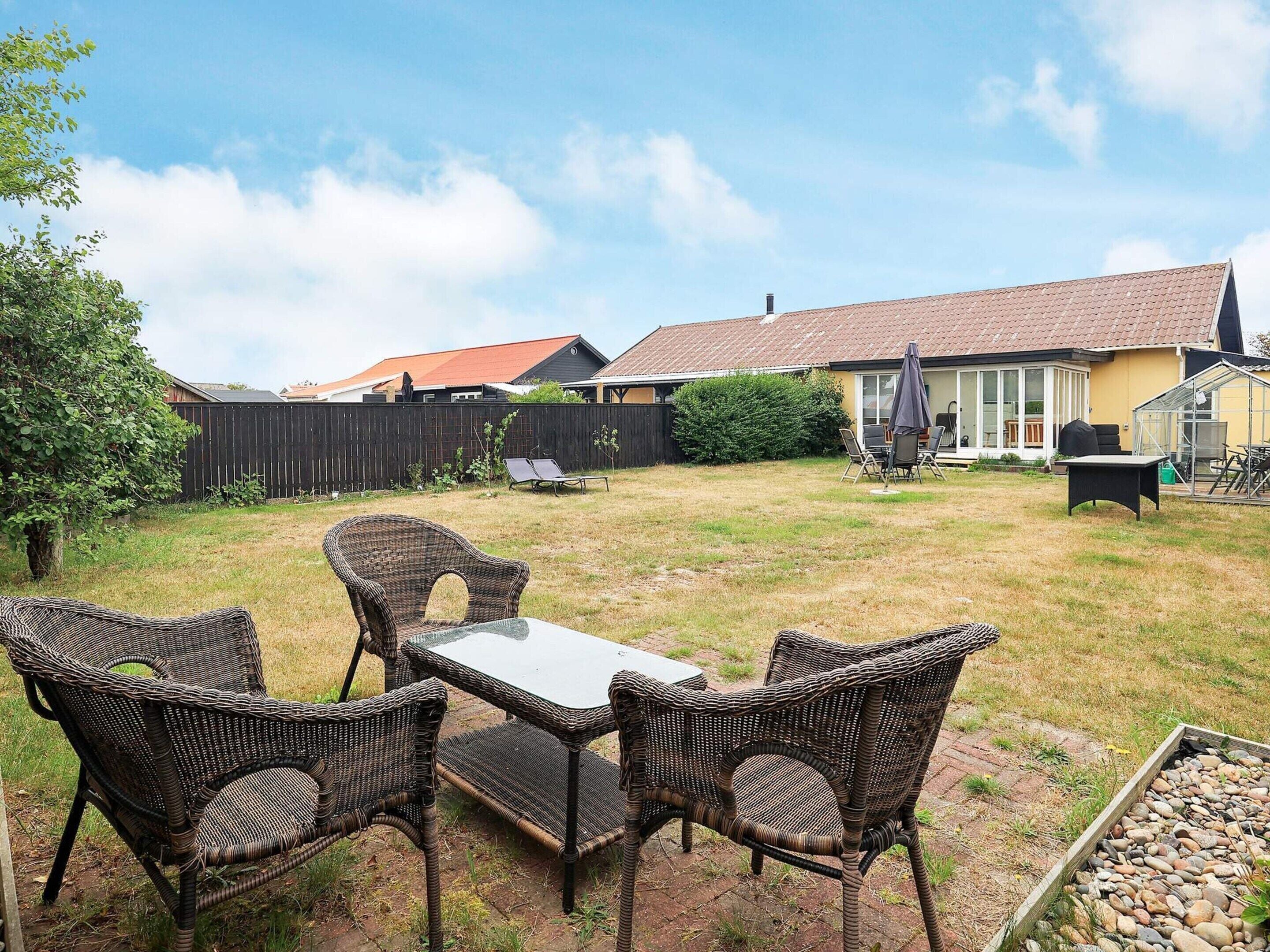 An outdoor patio with wicker furniture and a glass table, surrounded by a grassy yard and a wooden fence.
