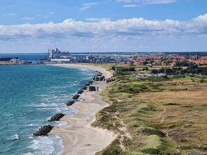 Zona costera con una playa arenosa, un rompeolas rocoso y un paisaje urbano distante con edificios y un puente.