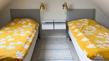 A small, single-room accommodation with two beds, each with a yellow floral bedspread and a grey headboard. There are two bedside tables with lamps, a small white dresser, and a grey rug on the floor.