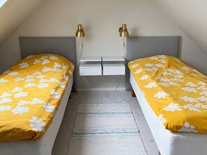 A small, single-room accommodation with two beds, each with a yellow floral bedspread and a grey headboard. There are two bedside tables with lamps, a small white dresser, and a grey rug on the floor.
