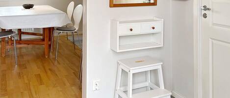 Une pièce avec une table blanche, des chaises, un petit tabouret blanc, une étagère fixée au mur et une porte.