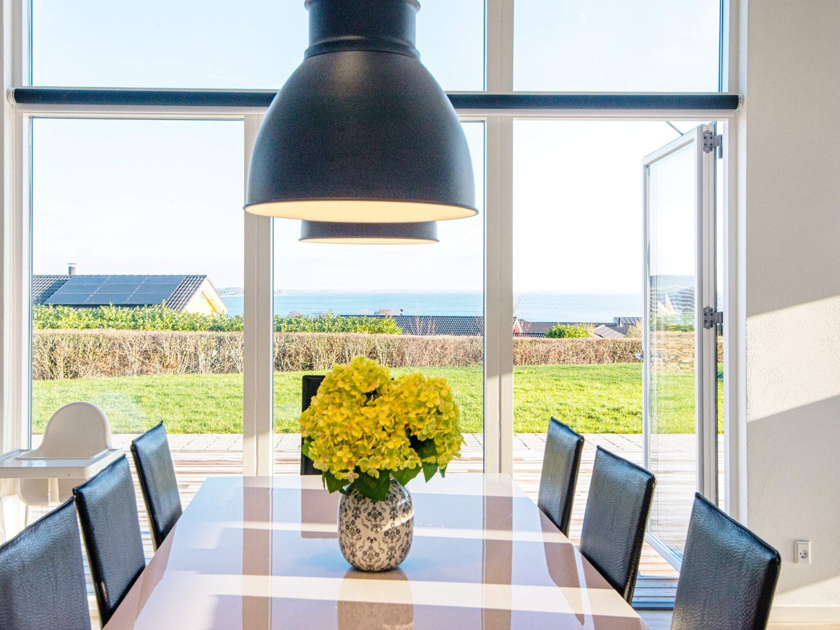 A dining table with chairs, a vase of yellow flowers, and a view of the sea through large windows.