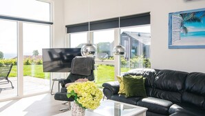 A modern living room with a black leather sofa, a white coffee table, and a glass-top table with a vase of flowers. There is a large window with a view of a house and greenery outside. - 10 Person Holiday Home in Sjolund (Sjølund)
