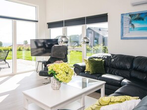 Ein modernes Wohnzimmer mit einer schwarzen Ledersofa, einem weißen Couchtisch und einem Glastisch mit Blumenvase. Ein großes Fenster bietet Blick auf ein Haus und Grünflächen.