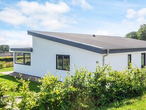 A modern single-story house with a grey roof and white exterior walls, surrounded by lush greenery.